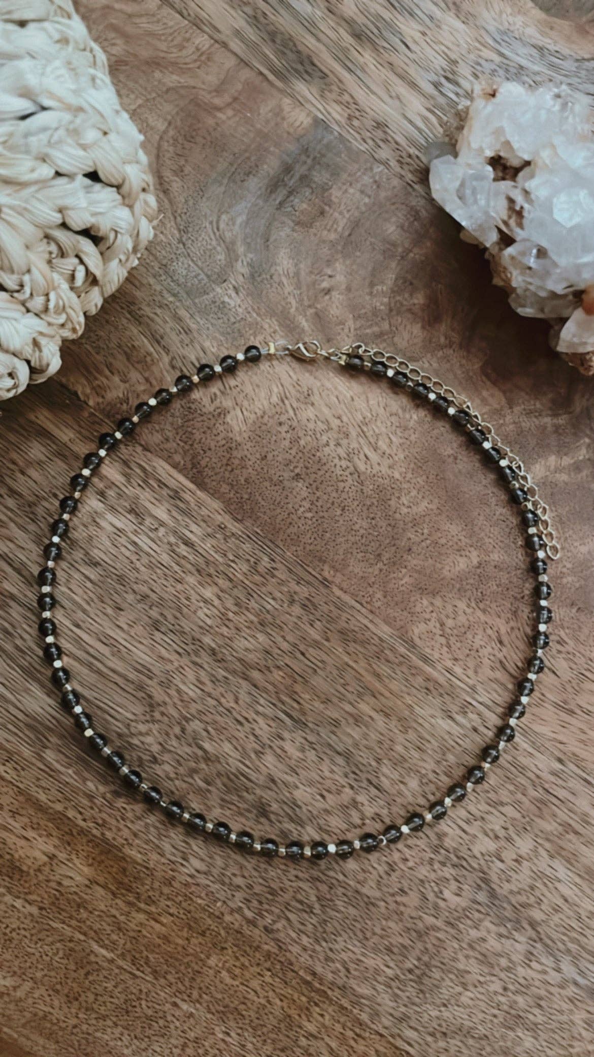 A Smoky Quartz Choker featuring smoky quartz beads and gold accent beads, displayed on a wooden surface with crystal decor.