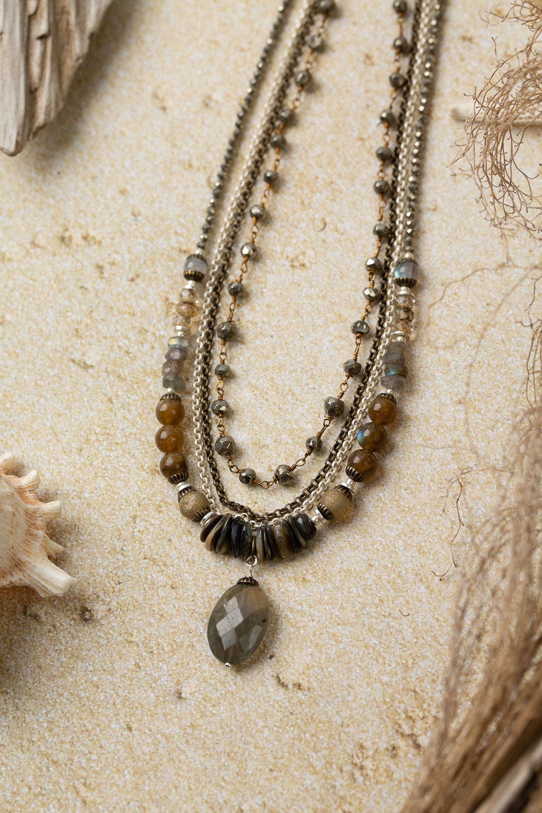 Handcrafted Dunes Collection Multistrand Necklace laid on a sandy surface, showcasing the elegant combination of Czech glass, abalone, and labradorite gemstones with a sterling silver chain.