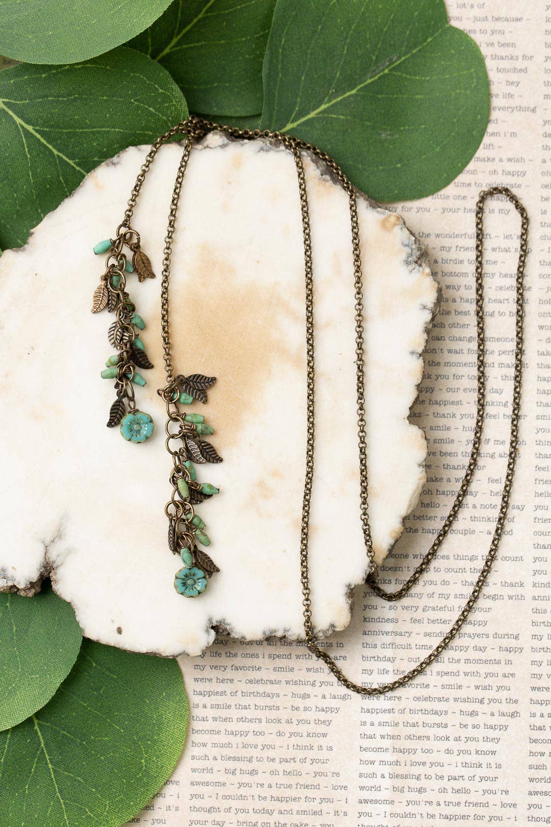 A Turquoise and Czech Glass Lariat Necklace featuring antique brass and leaf charms, displayed on a white marble background with green leaves.