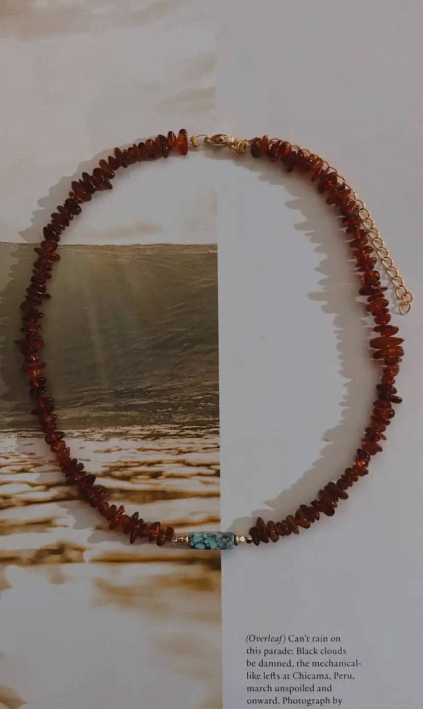 A Slow Burn Choker featuring amber beads and a turquoise tube bead, displayed on a light background with a scenic backdrop.