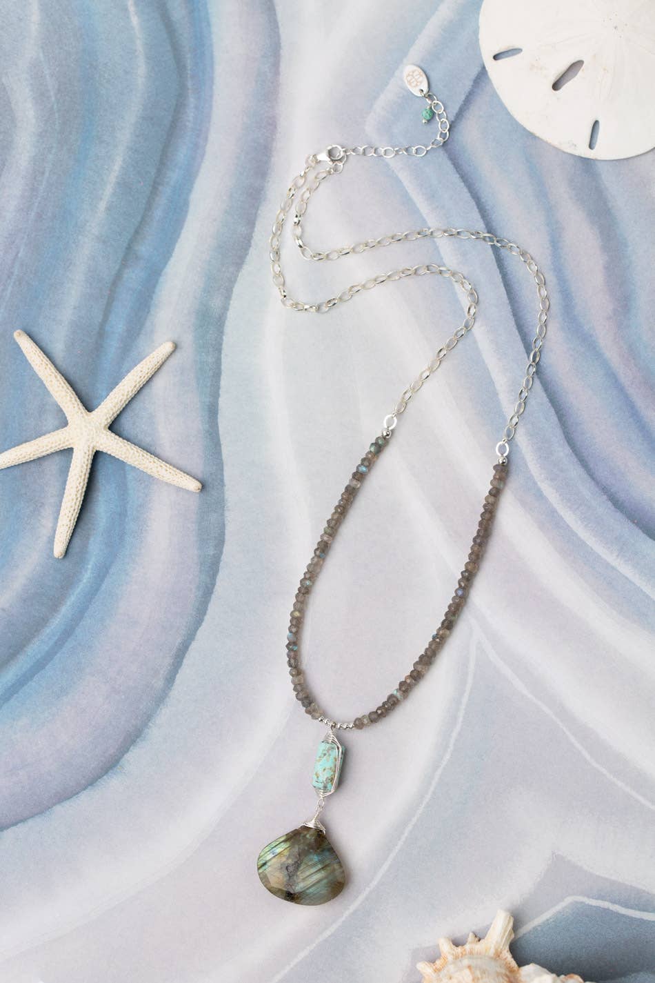 A close-up of a sterling silver necklace from the Mystic Collection, featuring labradorite and turquoise pendants, laid on a serene, wavy blue background with a starfish and sand dollar.